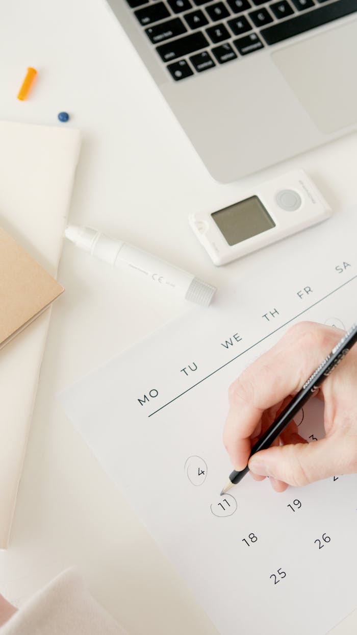 A person marking dates on a calendar, with a glucometer and laptop nearby, suggesting diabetes monitoring.